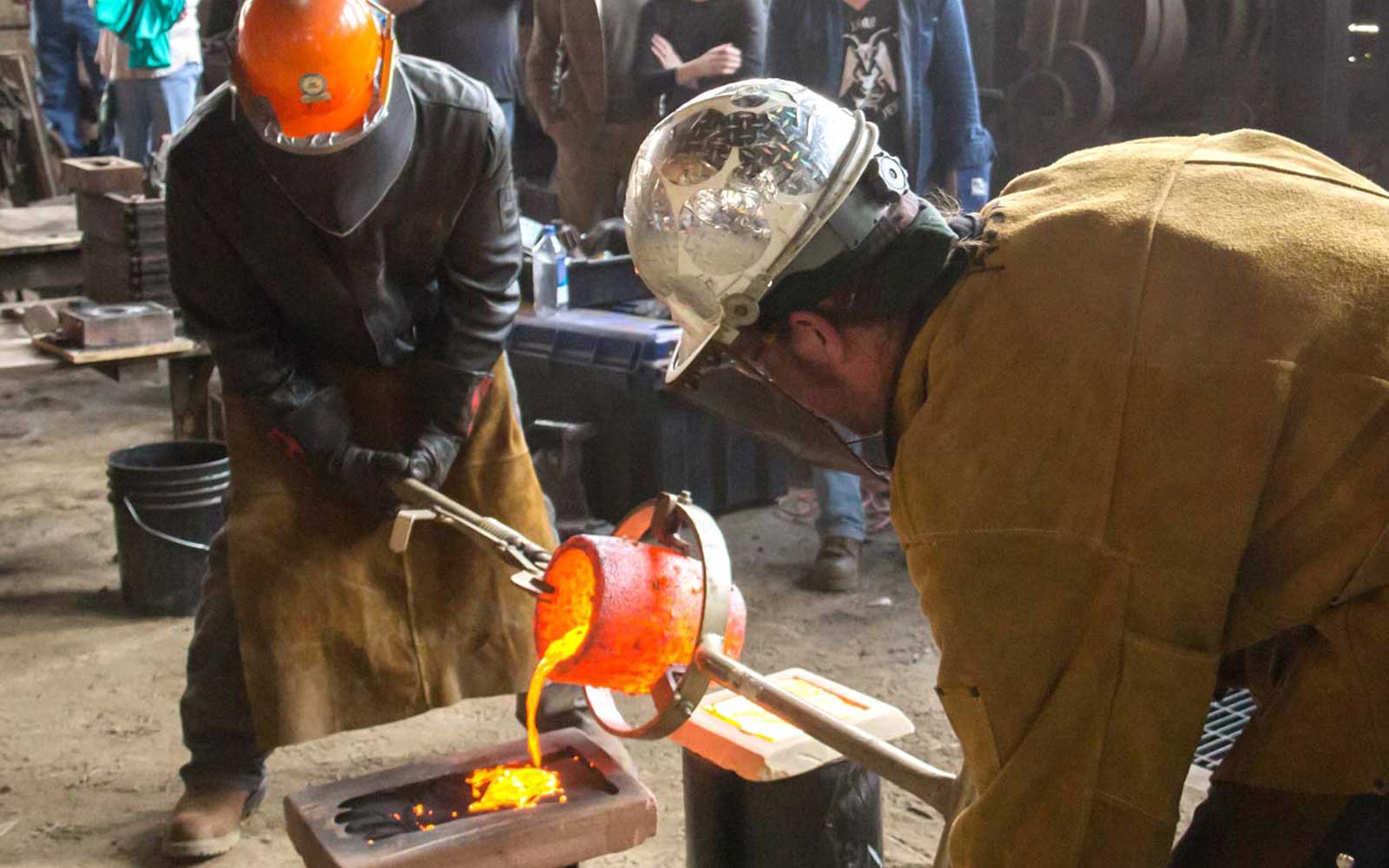 Two men wearing hard hats pouring molten iron.