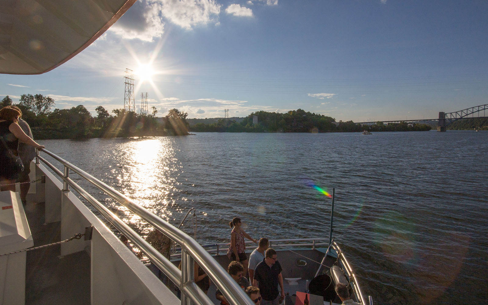 A view of the Ohio River that includes a late afternoon sun and people on the bow of the boat.