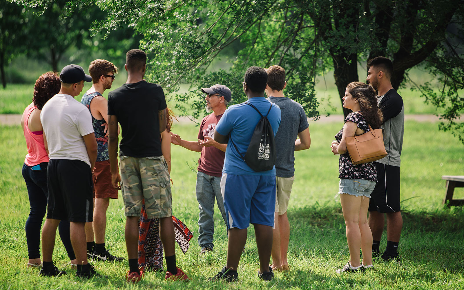 A group of young adults surround a tour guide in the shade of a tree.