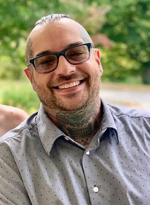 A smiling, 40ish, white man with visible tattoos wearing black rim glasses and a gray button down shirt,
