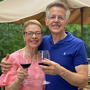 A middle aged white couple holding up wine glasses. He wears a blue polo shirt and she a pink flutter sleeve top.