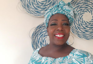 A smiling black woman dressed in a beautiful blue and white print with a matching headwrap with blue and white traditional baskets in the background.