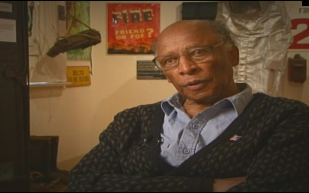 A Black man in a button down shirt and sweater, sitting in a museum.