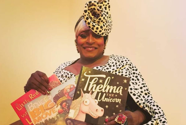 A Black drag queen poses with children's books while wearing a animal print dress and a matching fancy hat.