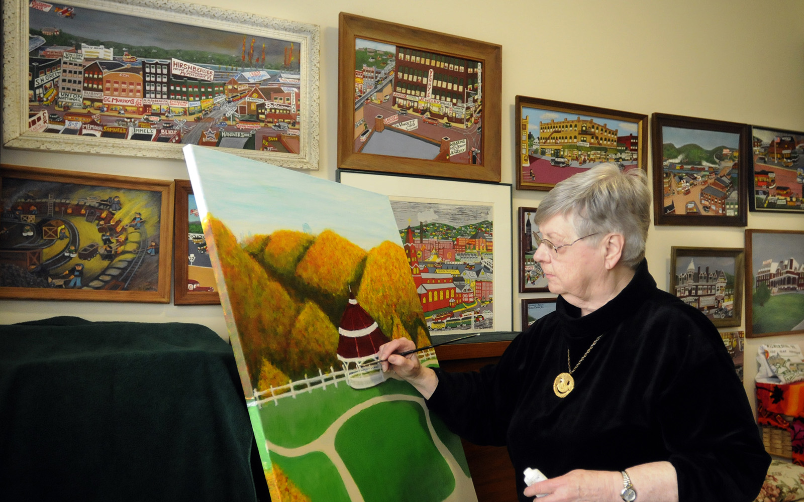 A gray haired woman in a black sweater paints a pastoral scene. Behind her is a wall of her other paintings.
