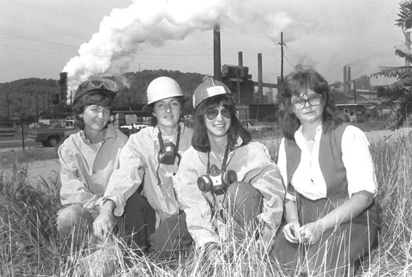 A black and white image of four white women, two in hard hats and respirators in front of a smoking steel mill.
