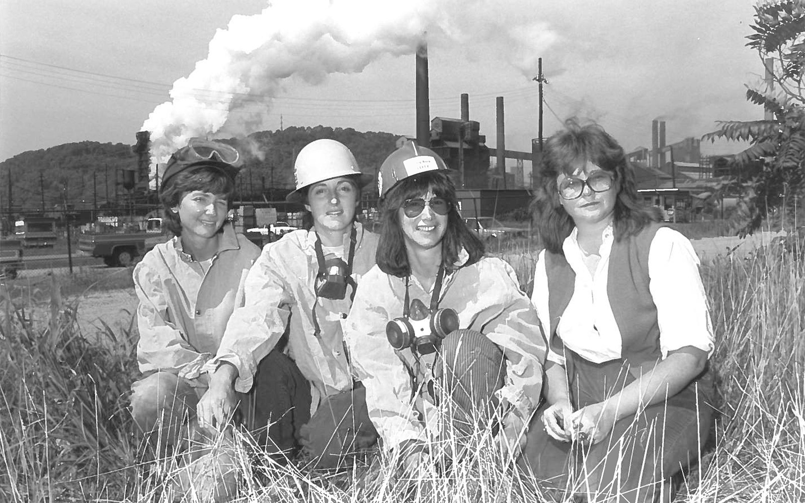 A black and white image of four white women, two in hard hats and respirators in front of a smoking steel mill.