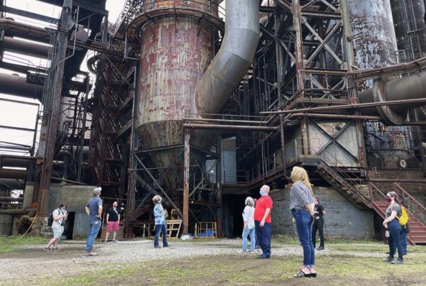 A small group of people stand in courtyard, looking up at the Carrie Blast Furnaces.