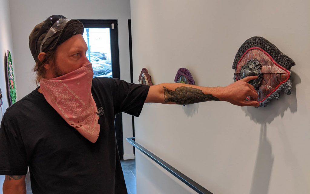 A white man wearing a faded red bandana mask points at the iris in his sculpture of an eye.