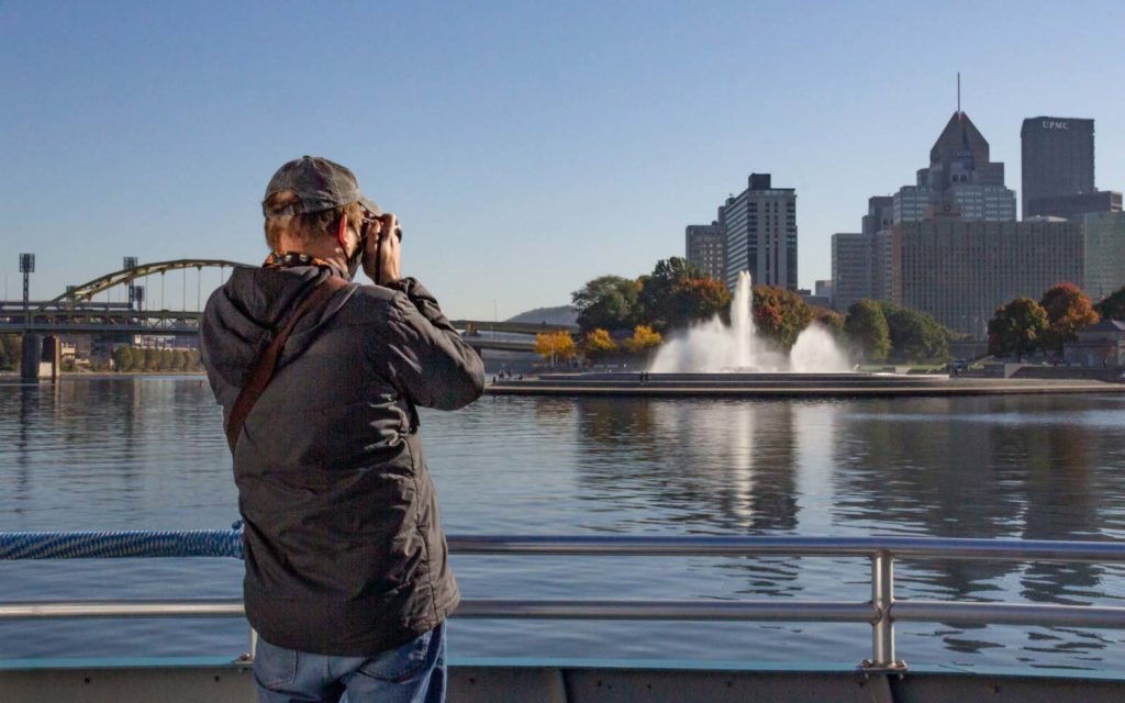 A white male in a ball cap photographs the Pittsburgh skyline from the riverboat.