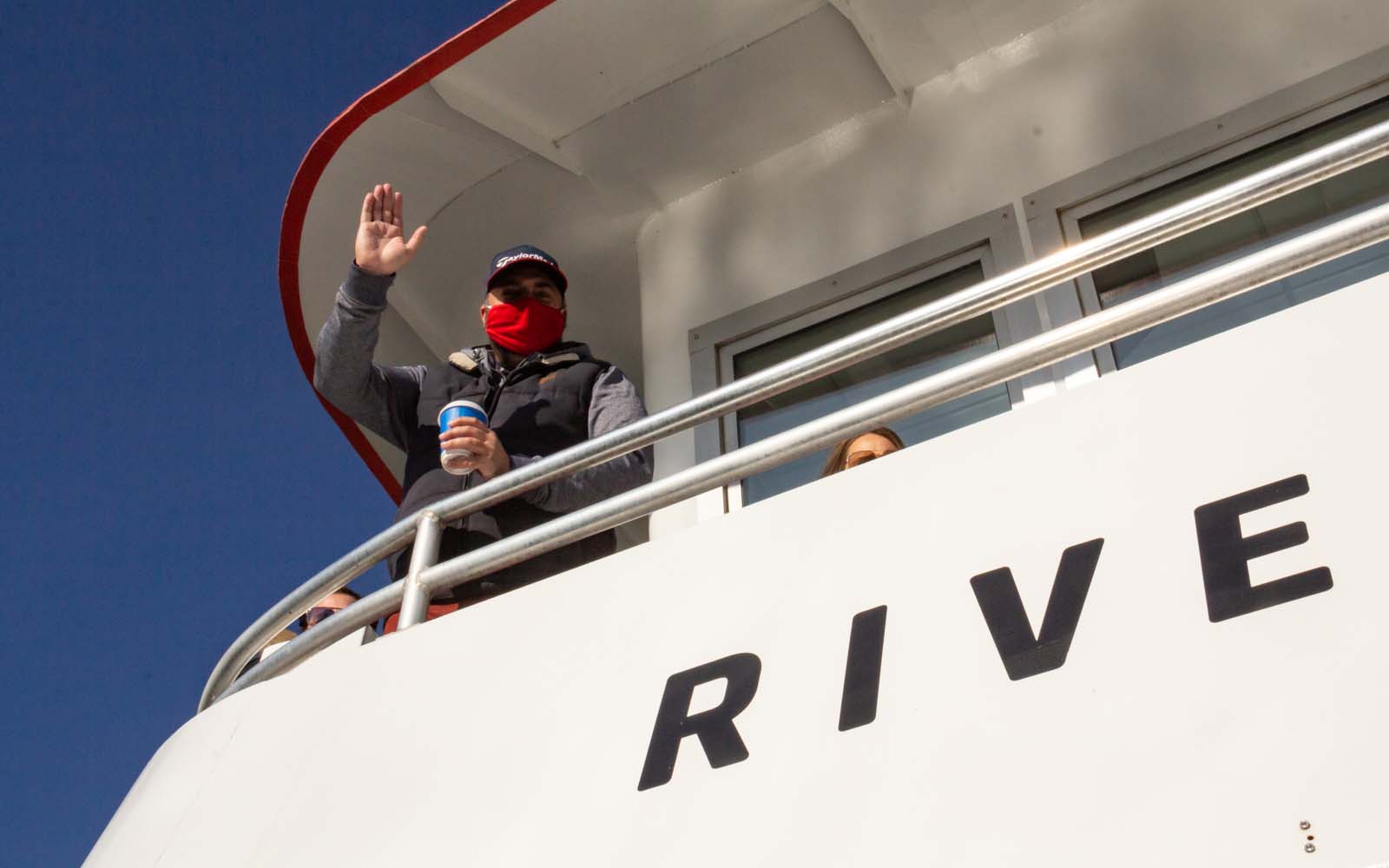 A man in a ball cap and a red mask waves at the camera from the upper deck of the boat.
