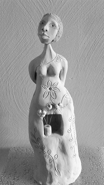 A porcelain figure with her hands behind her back, flower on her dress and a small box sculpted out between her hips that holds a vase with flowers inside