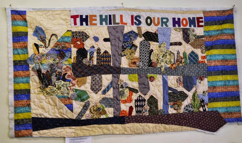 A quilt with a white background, stripes on the left & right border and a colorful patchwork in the middle that reads "The Hill is our home."