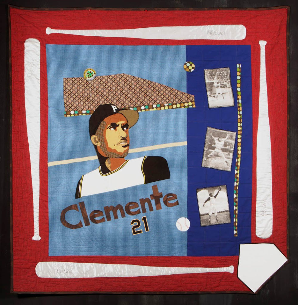 A quilt with a red border with a white baseball bat on each side that has blue fabric in the middle and a portrait in fabric of Roberto Clemente. It also has photographs of him on fabric, along with his name and number "21".