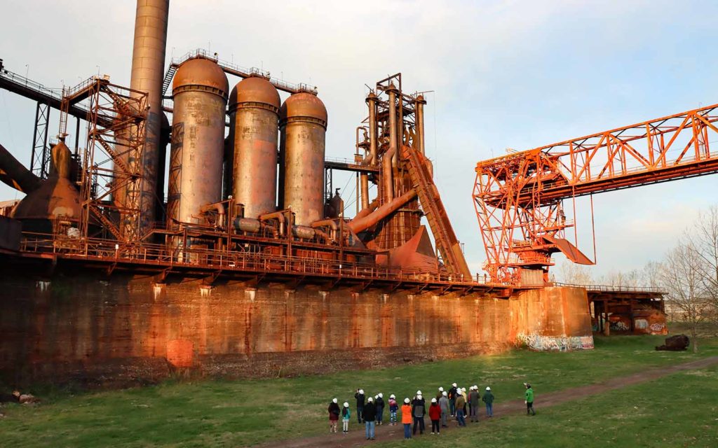 A group in hard hat walk through the ore yard in front of the Carrie Blast Furnaces.