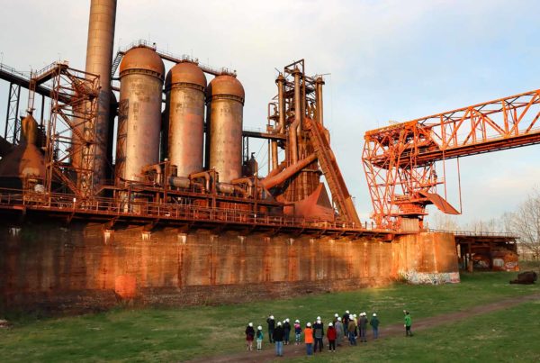 A group in hard hat walk through the ore yard in front of the Carrie Blast Furnaces.