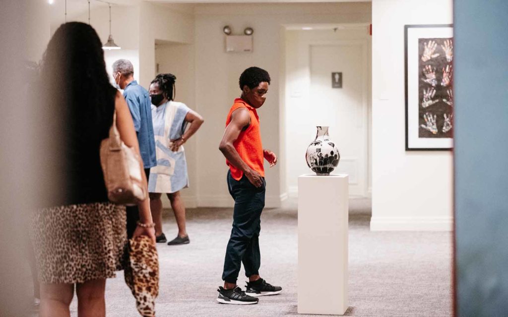 Four people of color walk through a white walled gallery showing paintings and sculpture.