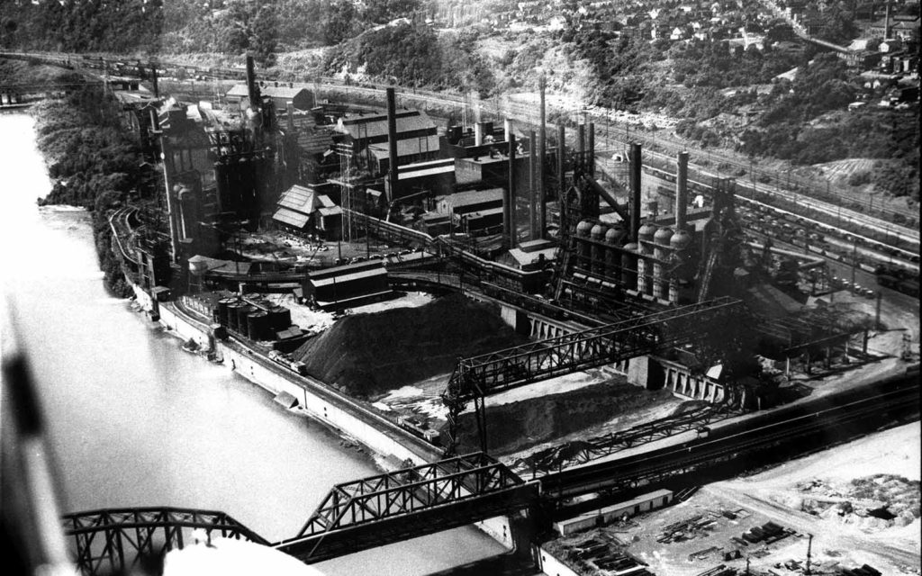 A black and white aerial photo of the Carrie Furnace site when it fully active, showing lots of buildings, smoke, and raw materials.