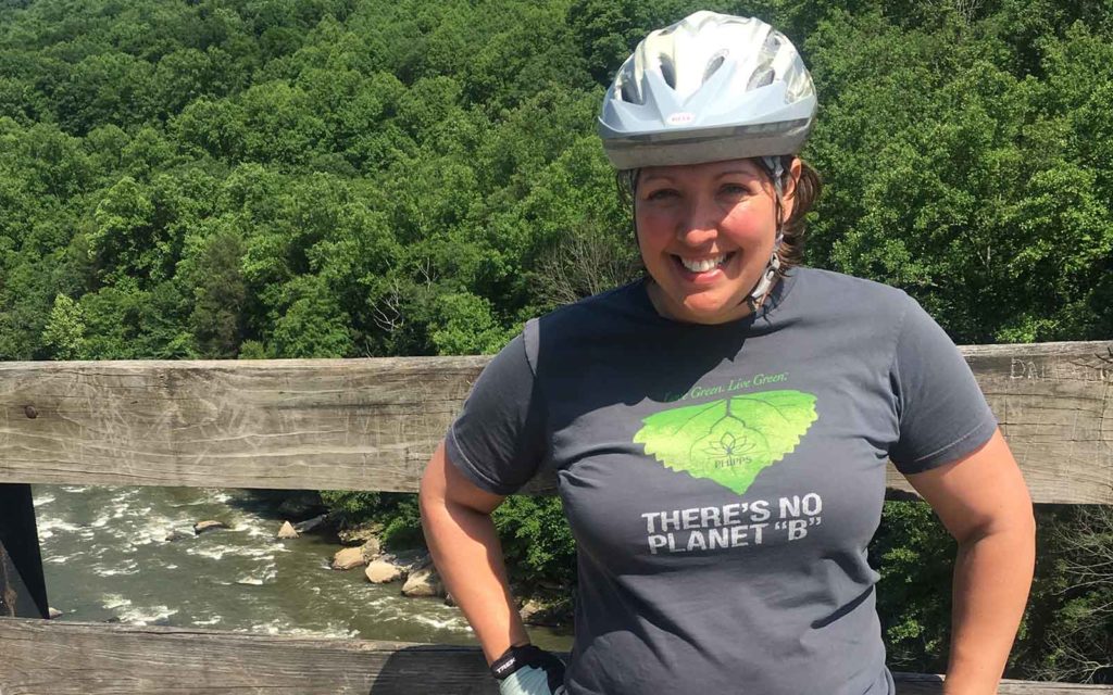 A woman in a gray t-shirt and bike helmet poses for a photo on a bridge above a river showing whitewater caps.