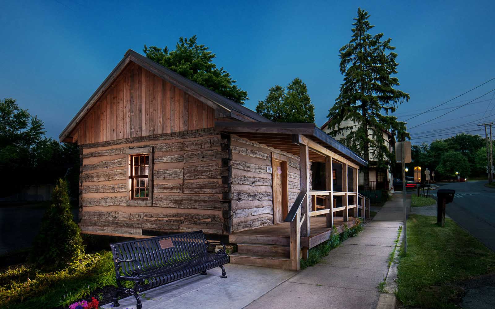 A log cabin with a sidewalk in front and a large pine tree to the side.