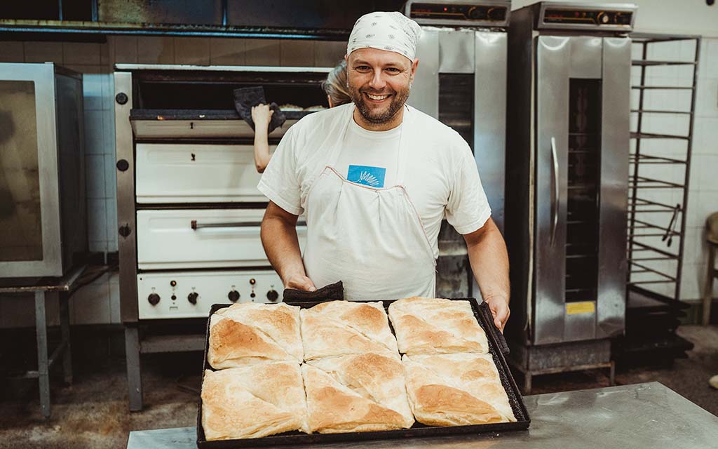 A baker in white displays a pan of bread.