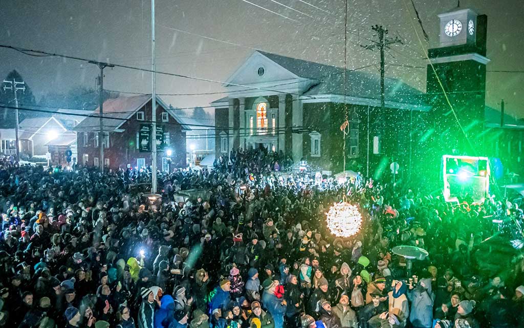 Hundreds gather around a well lit new years ball as snow falls in front of historic, columned buildings and a clocktower.