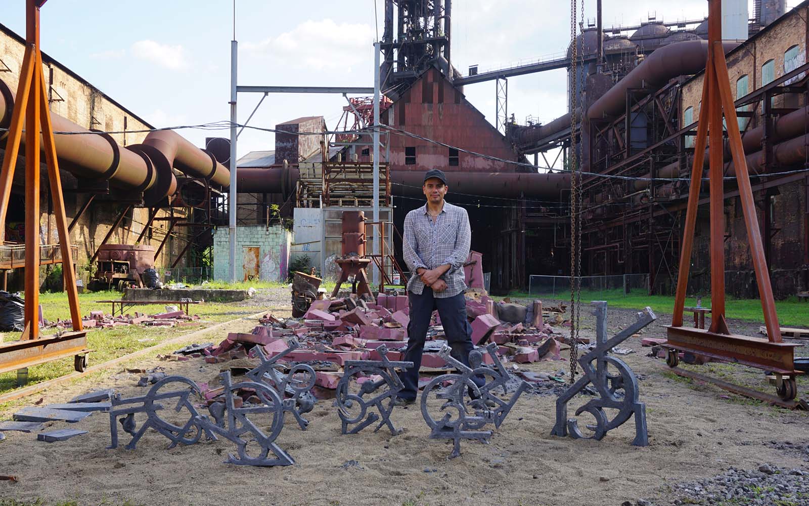 a man stands with sculptures in front of a small scale iron furnaces with an industrail former blast furnace behind them