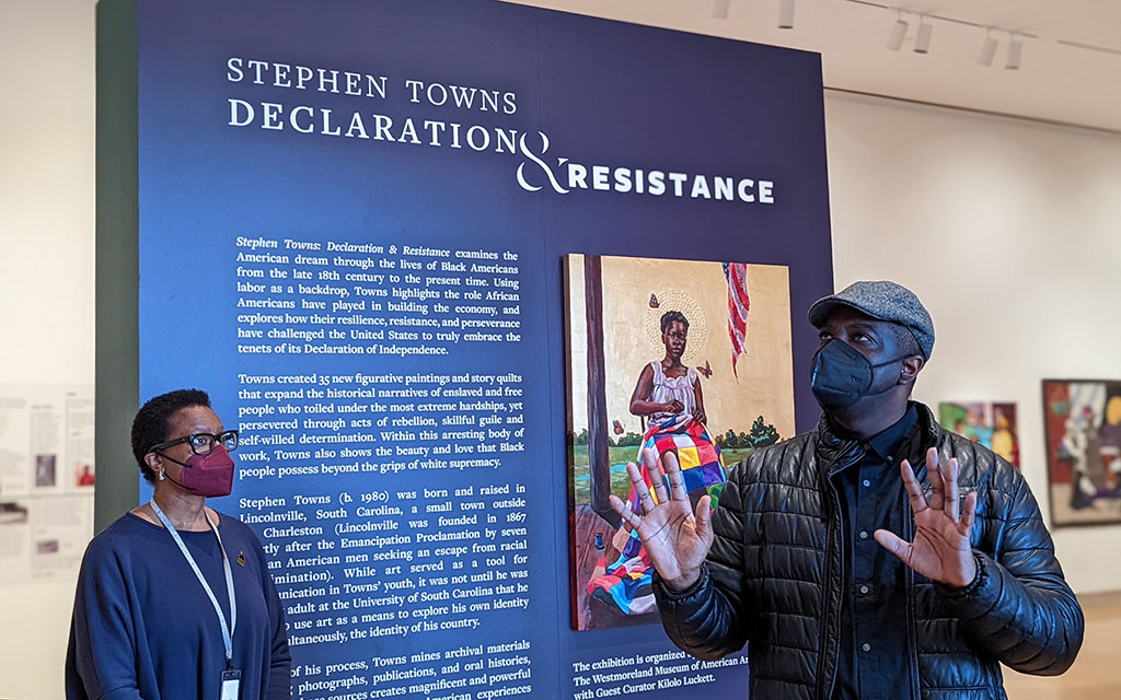 A Black woman and A black man stand in front of the wall with text labeling the exhibition. He is holding his hands up as he speaks.