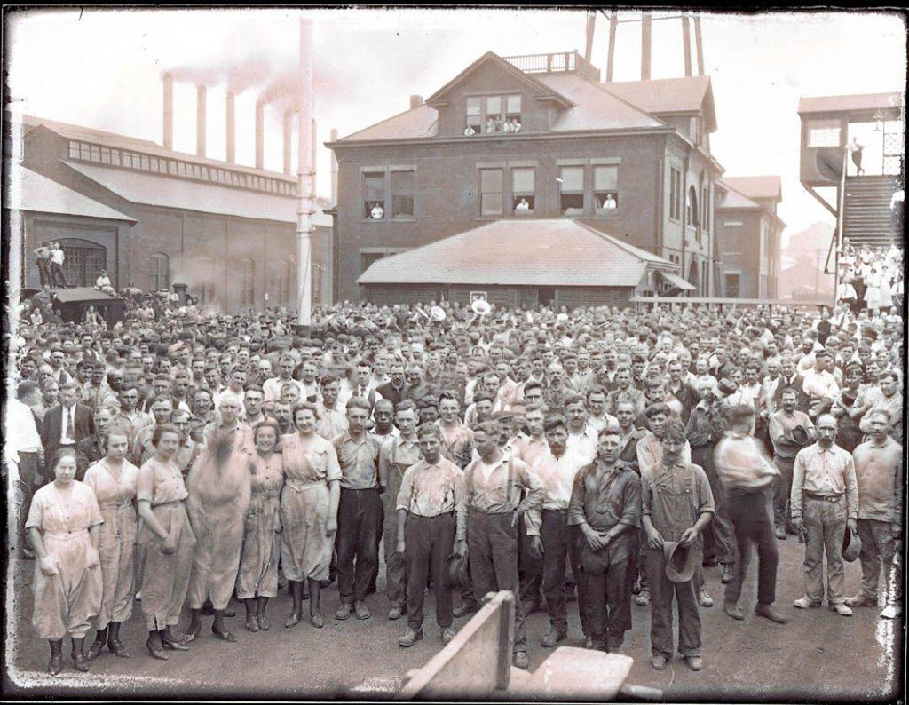 Hundreds of workers, mostly whilte and nearly all in dirtied work clothes, pose for a picture in front of what may be an administrative building with a shed full of smokestacks adjacent to it.