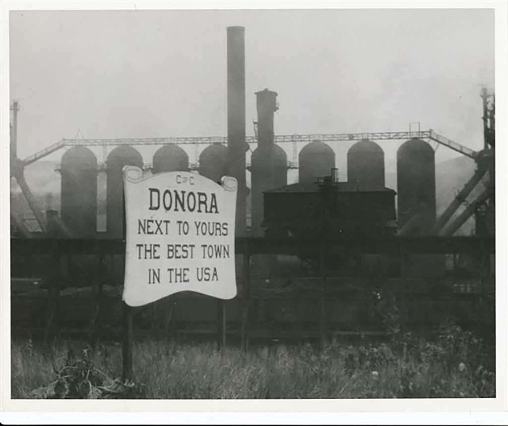 Eight blast furnace stoves and one stack rise up behind a sign that reads "Donora Next to Yours the Best Town in the USA."
