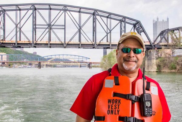 A black man wearing a work life vest on a boat with a bridge the skyline in the background.