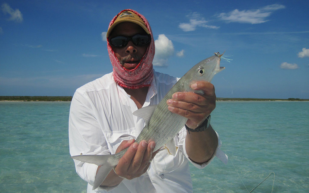 Blue skies and clear pale green water surround John, dressed in a white shirt with his head covered in a red cloth, as he holds up a nearly clear fish.
