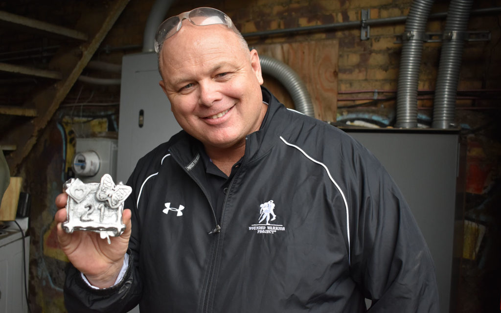A bald white man in a black jacket with the wounded warriors project logo holds up his aluminum casting.