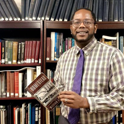 A black man in a plaid button down shirt with tie in front of books.
