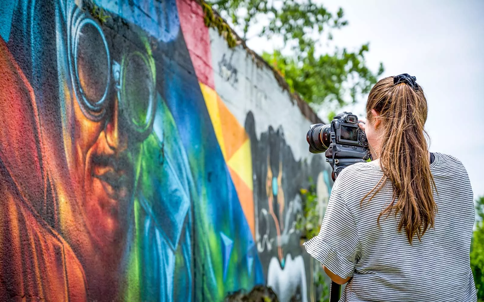 A woman with a long brown ponytail in a t-shirt takes a photo of a colorful mural.