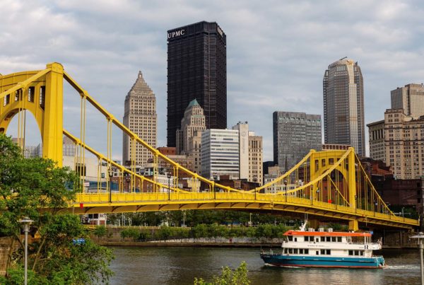 The riverboat is passing under a bridge with the Pittsburgh skyline.