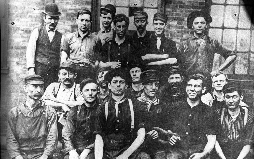 Rugged looking men, some smiling, pose for the camera, all wearing workwear, with the exception of one guy in a vest and bowler hat.