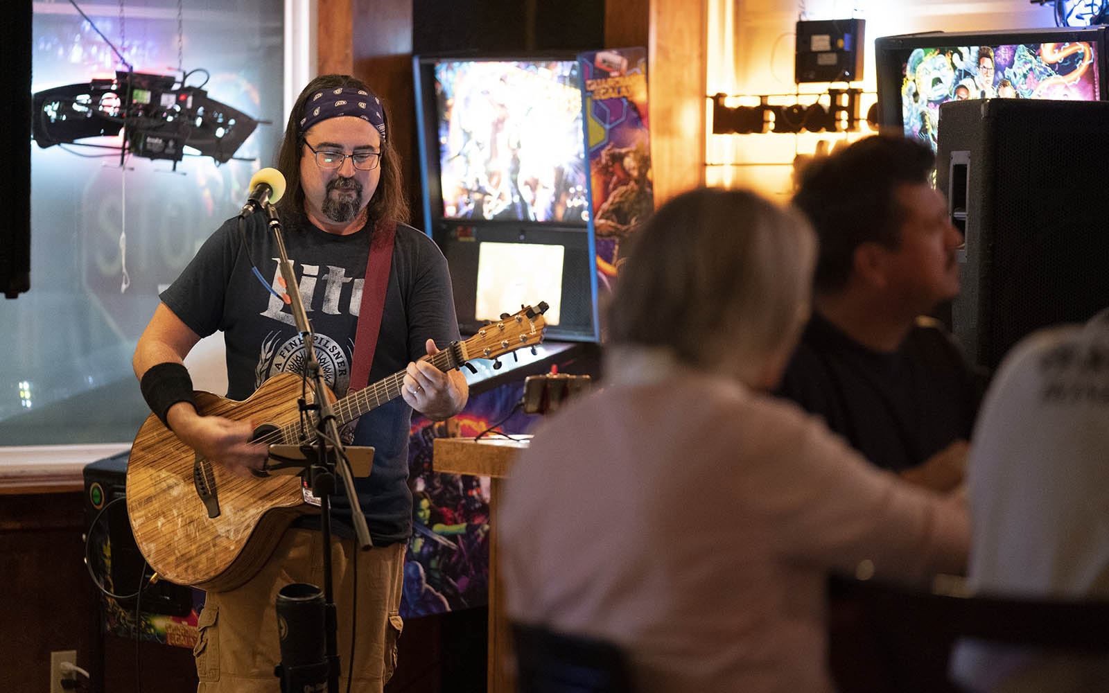 A white man with a goatee, glasses, and a bandana over his long hair plays an acoustic guitar standing in front of the glass window of a bar front with patrons gathered in the foreground.