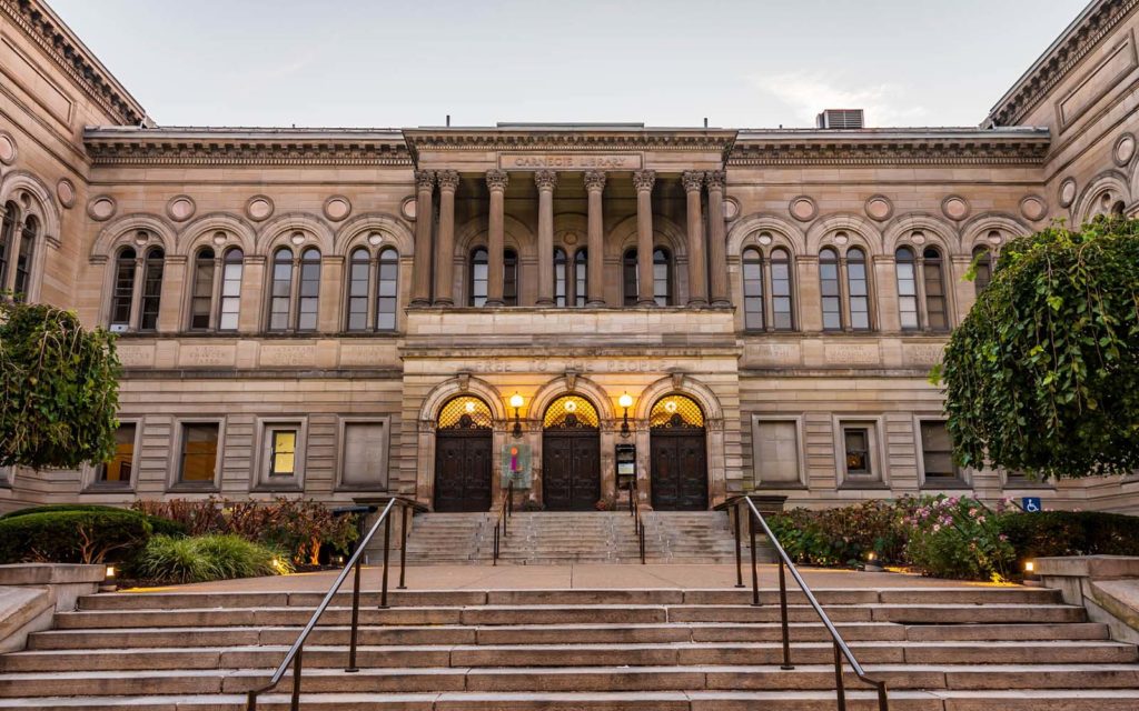 A marble and granite structure in the Beaux-Arts style, two stories with a promenade of steps leading to three arching doorways underneath a covered balcony supported by Greco-Roman columns..