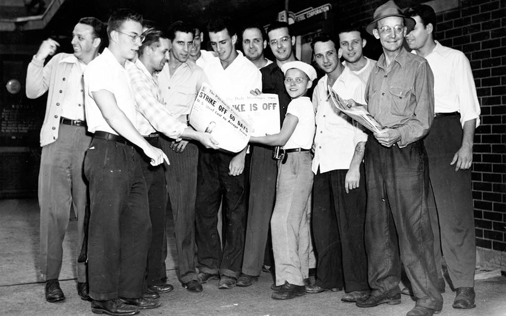 About 15 men in dark pants and white shirts joyfully gather with newspapers showing headlines about the end of a strike.