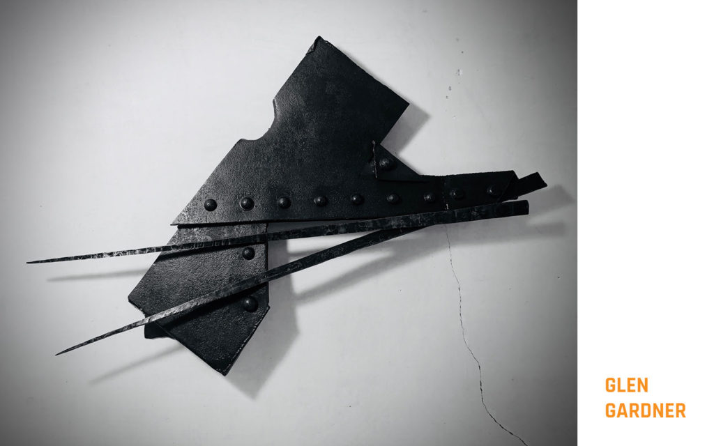 A dark-colored irregular shaped piece of metal with two arrowlike protrusions extending from it hanging on a white wall.