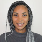 A youthful brown skinned woman with silver and black braids, smiling in a gray mock turtleneck.