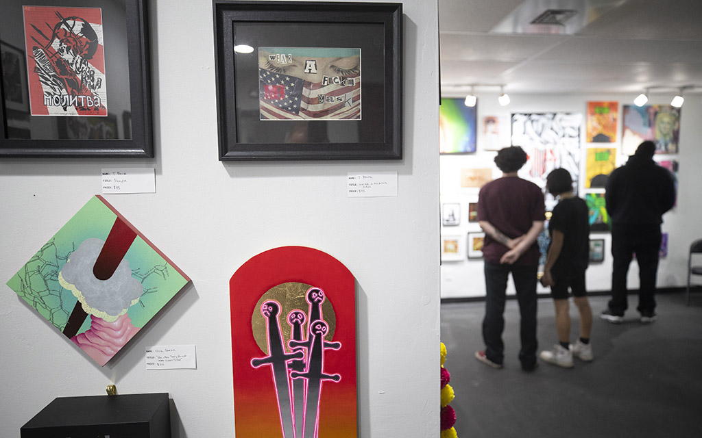 In left foreground, artworks hang on the wall. To the right a crowd gathers around other colorful artworks.