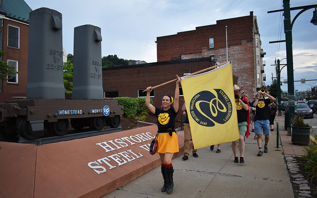 A woman in an gold skirt and black top waves and black and gold flag and leads a troupe of traveling musicians.