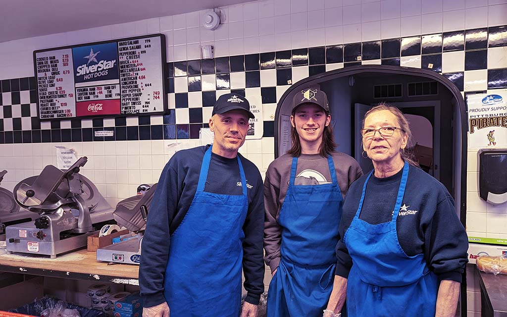 Three employees in blue aprons stand in front of a white tile wall with a black and white checkered tile border near the ceiling. A meat slicer is visible on the counter behind them.