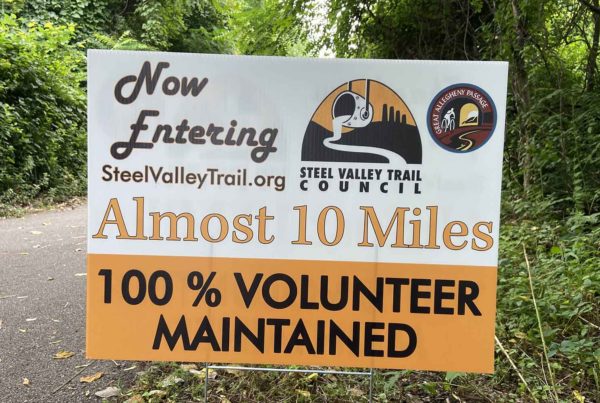 A sign along a trail reads "Now entering Steel Valley Trail dot org, almost 10 miles, 100% volunteer maintained"
