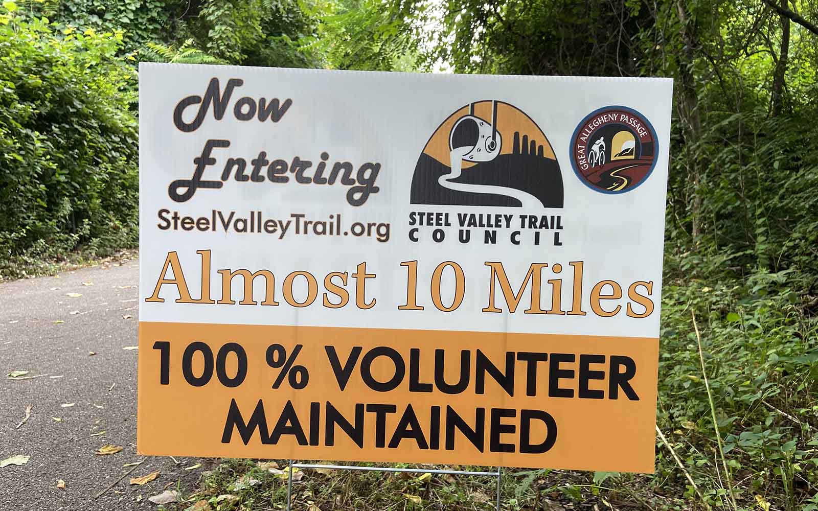 A sign along a trail reads "Now entering Steel Valley Trail dot org, almost 10 miles, 100% volunteer maintained"