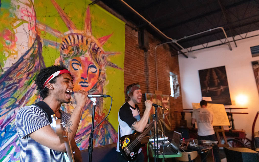 Two rocking musicians sing into microphones with colorful art behind them.