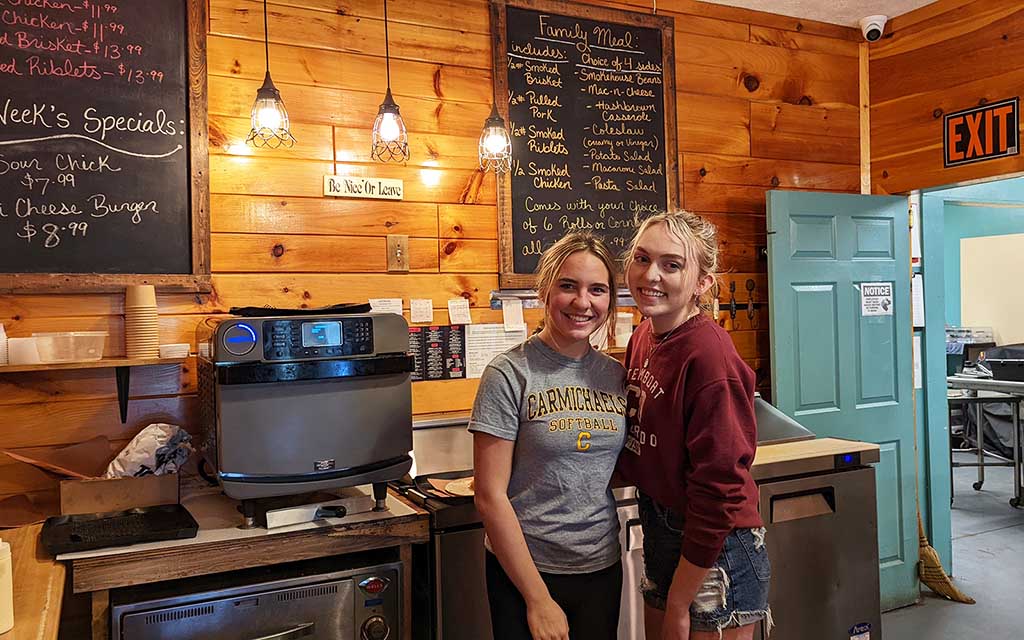 Two teen girls with pulled back sandy blond hair smile for the camera in front of a horizontal wood paneled wall with special written on chalk boards mounted to it.