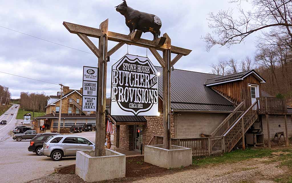 A carved cow is perched above a large sign reading Stone House Butcher and Provisions, Circa 1822." The shop emerges from behind the sign and the road disappears over the ridge on the left side of the frame.